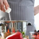 Solenie jedla pri varení — nerezový hrniec s čerstvou zeleninou na kuchynskej doske