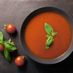Krémová paradajková polievka s čerstvou bazalkou v tmavej miske s cherry paradajkami