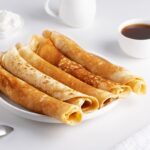 Tenké palacinky stočené do roliek na bielom tanieri s kávou a šľahačkou