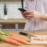 Žena hľadá recept na mobile pri krájaní mrkvy v kuchyni