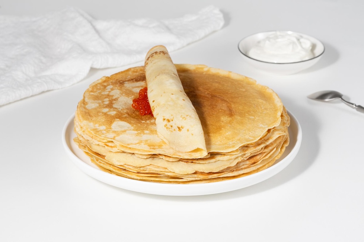 Stoh tenkých francúzskych crêpes na bielom tanieri s jednou zrolovanou palacinkou a kyslou smotanou
