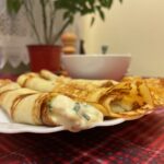 Maslo alebo olej na palacinky – čo dáva lepší výsledok? Zrolované tenké palacinky so syrovou plnkou na bielom tanieri na červenom obruse