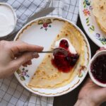 Ako urobiť palacinky dokonale tenké – 6 trikov Tenká palacinka s lesným džemom a smotanou na kvetovanom tanieri, v pozadí stoh palaciniek